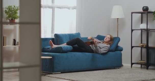 Romantyczna młoda dziewczyna w szarej bluzie gra na gitarze w domu. Kobieta gitarzysta spoczywa na niebieskiej kanapie spogląda w górę. Mało prawdopodobne.. — Wideo stockowe