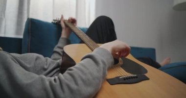 Sarı saçlı genç bir kızın beyaz bir oturma odasındaki kanepede uzanmış görüntüsü. O, akustik bir gitarda rahatlatıcı müzik çalar. Yakın plan..