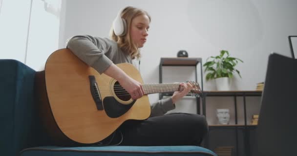 Het meisje luistert naar muziek in een witte koptelefoon met een laptop. Een vrouw zit op een blauwe bank in een lichte woonkamer. De muzikant kiest een melodie voor de gitaar. Tweede middelmatig schot. — Stockvideo