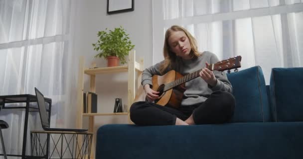 Fiatal gyönyörű lány ül egy puha kék kanapén lótuszpózban a nappaliban. Egy tinédzser megtanul gitározni egy laptoppal egyedül. Földi szöglövés — Stock videók