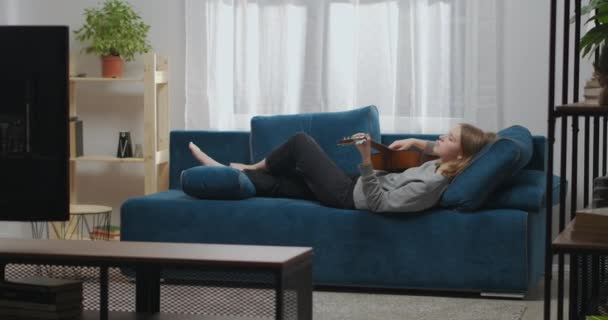 Side view of romantic young girl in gray sweatshirt playing guitar at home. Female guitarist resting on a blue sofa. Long shot. — Vídeos de Stock