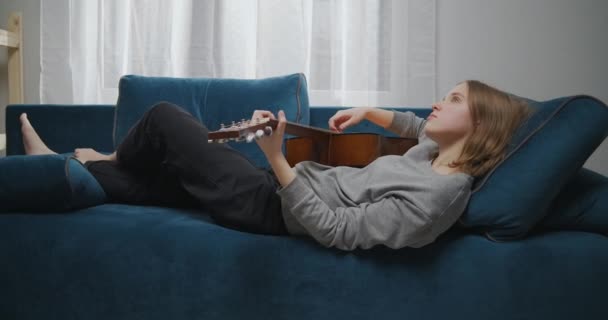 Vista lateral de un músico suelto con el pelo rubio acostado en un sofá en casa en un fin de semana. Girl está disfrutando tocando una alegre melodía en una guitarra acústica. El músico sacude la pierna y la cabeza con tanta melodía. — Vídeos de Stock