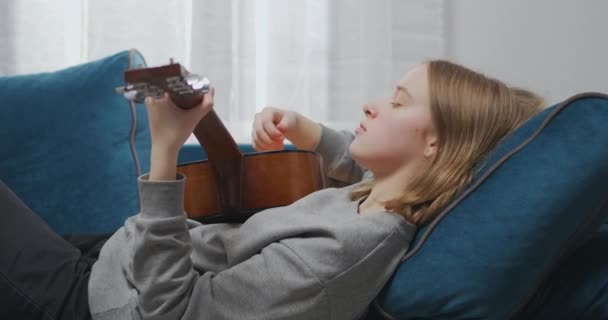 Atraktivní dívka v domácím oblečení hraje na kytaru v obývacím pokoji, relaxaci na měkké pohovce v odpoledních hodinách.