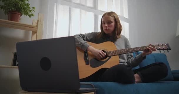 Fiatal gyönyörű lány ül egy puha kék kanapén lótuszpózban a nappaliban. A tinédzser mesterek hangszeren játszanak. Egy zenész megtanul gitározni egy laptoppal egyedül.. — Stock videók