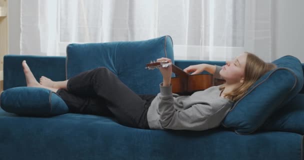 Sisi tampilan gadis santai bermain gitar di ruang tamu. Wanita impian berbaring di sofa dengan alat musik. Gitaris perempuan beristirahat di sofa. Wanita yang menarik memainkan musik lambat pada gitar akustik — Stok Video