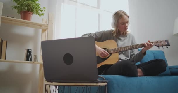 Dziewczyna słucha muzyki na słuchawkach za pomocą laptopa. Kobieta siedzi na niebieskiej kanapie w jasnym salonie. Muzyk wybiera melodię na gitarę. — Wideo stockowe