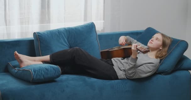 Widok z boku spokojnej kobiety grającej na gitarze w salonie. Romantyczna młoda dziewczyna leżąca na kanapie z instrumentem muzycznym. Kobieta gitarzysta spoczywa na miękkiej sofie. — Wideo stockowe