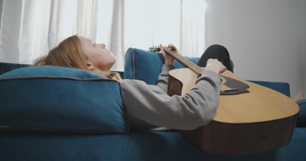 Side view of a young girl with blond hair lies on a sofa in a white living room. She plays relaxing music on an acoustic guitar. Medium shot — Stock Video
