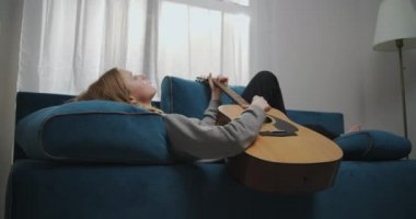 Beyaz bir oturma odasındaki yumuşak bir kanepede uzanmış sarı saçlı genç bir kızın doğru görüntüsü. O, rahatlatıcı klasik gitar müziği çalar. İlk ortam planı.