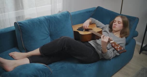 Chica joven con ropa casual se encuentra descalza en el sofá en su apartamento. Toca una melodía relajante en una guitarra acústica. — Vídeos de Stock