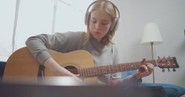 Bir kız, dizüstü bilgisayarında dinleme cihazı olan büyük beyaz kulaklıklarla müzik dersi dinliyor. Genç, gitar çalmanın temellerini özenle öğreniyor. Pencereden gelen güneş ışığı.