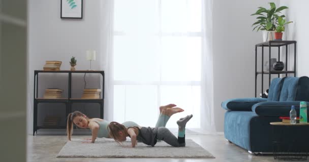 Entrenamiento matutino de madre e hija pequeña en la sala de estar, haciendo flexiones juntas, apoyadas en el suelo por las manos — Vídeos de Stock