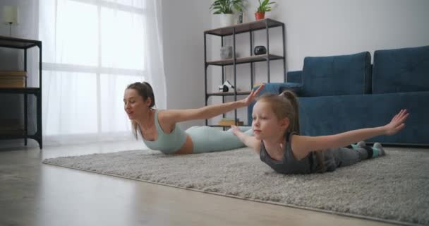 Mladá žena a její holčička spolu trénují v bytě, leží na podlaze a stoupá trup, zábava a zábava — Stock video
