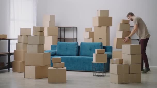 Junger Mann bewegt sich in Kartons. Junger Kater zieht in neue Wohnung Ein Mann sitzt nach seinem Einzug auf einem Sofa in einer neuen Wohnung — Stockvideo