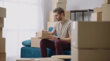 Genç bir adam bir karton kutuyu açıyor ve taşındıktan sonra koltukta oturan eşyaları çıkarıyor. Yeni bir daire alıyorum.