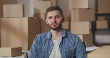 Küçük işletme sahibi sakallı yakışıklı genç adamın portresi kollarını kavuşturup gülümsüyor. Karton kutularla dolu bir depo..
