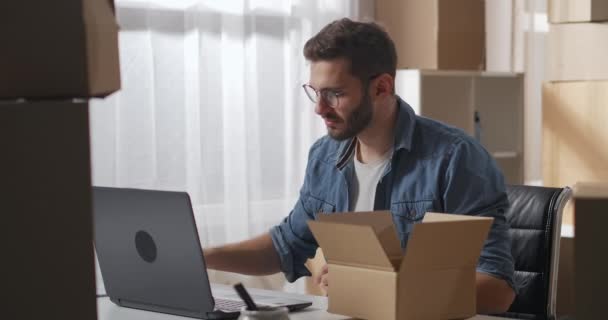 Ung manlig ägare med bärbar dator färdiga räkna objekt i pappkartong i lager. Mannen tog med de följande två lådorna.. — Stockvideo