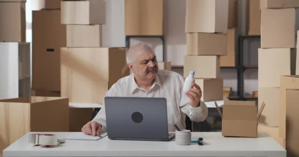 Mature man looks at bottle of household chemicals, checks the name of the product in the order with a ballpoint pen and paper. Trader packs the goods for shipment to the buyer. — Stock Video