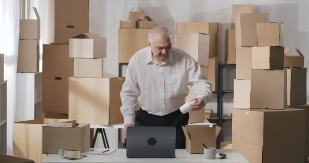 Businessman puts goods in a cardboard box indoors. A mature man checks the quantity of goods using a laptop and a clipboard. — Stock Video