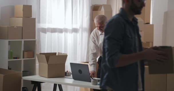 Business man puts VR glasses in a cardboard box indoors. Mature man is folding goods to be sent to customer. Long shot. — Stock Video