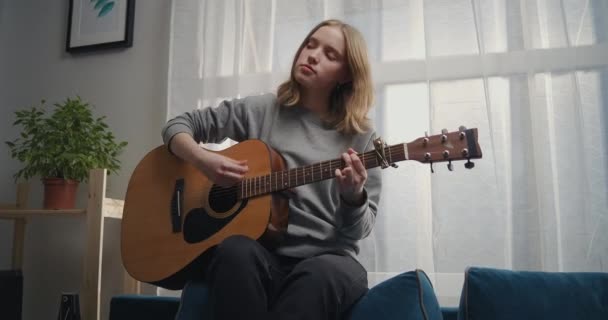 Jenta spiller gitar på sofaen. En kvinne alene skaper musikk i en hvit stue. Kunstneren spiller klassisk gitar. Musikeren komponerer en trist melodi. – stockvideo
