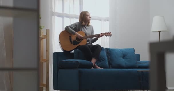 Un'adolescente con una felpa grigia e pantaloni neri suona una melodia calma con una chitarra acustica. Si siede sul retro di un divano blu a piedi nudi. Riprese in movimento. — Video Stock