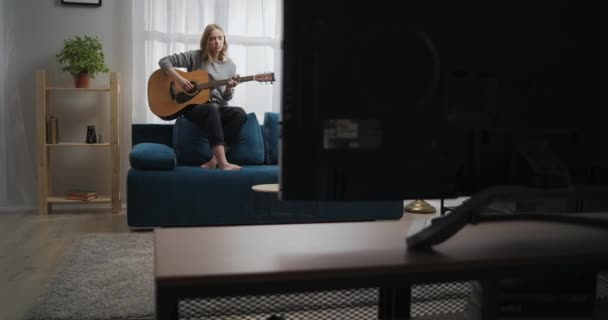 Gadis itu memainkan gitar di bagian belakang sofa. Seorang wanita menggubah musik di ruang tamu yang cerah dengan cahaya yang indah. Artis memainkan gitar klasik. Long shot in motion. — Stok Video