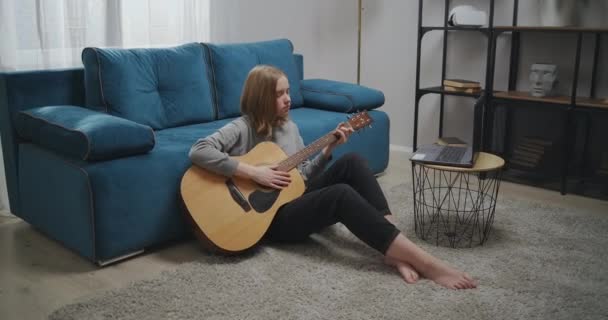 Nő néz online leckét, miközben ül a padlón a szobában. A lány elszigetelten tanul gitározni. Egy törekvő zenész megtanul hangszeren játszani laptoppal. Fejlövés — Stock videók