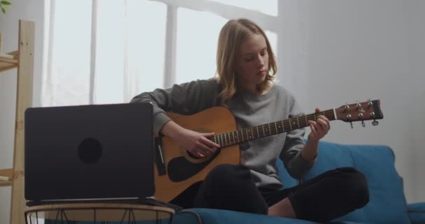 Mooi meisje kijken naar een online les geïsoleerd. Een jonge vrouw die thuis gitaar speelt. Een aspirant-muzikant leert een muziekinstrument bespelen met een laptop. — Stockvideo