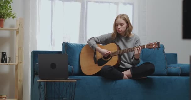 La adolescente se sienta en un sofá suave. El joven músico está aprendiendo a tocar la guitarra en el aislamiento. — Vídeo de stock