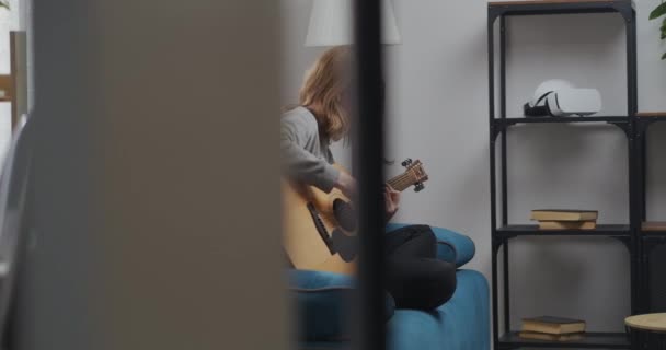 Un'adolescente con una felpa grigia suona musica tranquilla su una chitarra acustica nel suo tempo libero. — Video Stock
