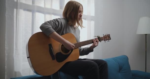 Het meisje speelt gitaar op de bank. Een vrouw alleen maakt trieste muziek in een witte woonkamer. De artiest speelt akoestische gitaar. De muzikant componeert een melodie. — Stockvideo
