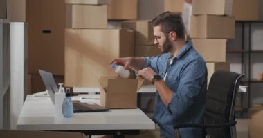 small business owner sits at a table in the office. male manager packs the vials of household chemicals one by one into cardboard box for delivery. First middle shot side view