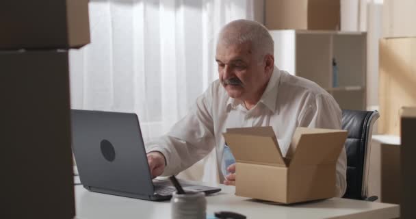 Propietario varón maduro en una camisa blanca con un ordenador portátil, contando artículos en una caja de cartón en un almacén. — Vídeo de stock