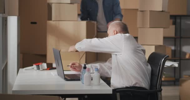 Boční pohled na malé podnikatele pracující společně ve skladu. Starý muž s laptopem, dává lahvičky domácích chemikálií do lepenkové krabice na stůl. — Stock video