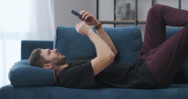 Yetişkin adam kanepede uzanıyor ve akıllı telefon kullanıyor, hafta sonu evde dinleniyor, piyango kazanıyor.