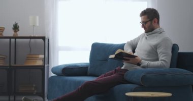Yetişkin adam evde kitap okuyor. Oturma odasındaki kanepede oturuyor. Hafta sonları dinleniyor.