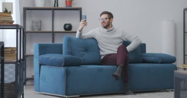 Adam evden görüntülü iletişim kuruyor, internet bağlantısı ve uygulaması olan akıllı telefon kullanıyor, el sallıyor ve merhaba diyor.