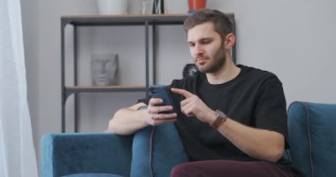 Genç adam akıllı telefonunda uygulamayı kullanıyor evde oturuyor, modern cihazların ekranını araklıyor ve video aracılığıyla insanlarla iletişim kuruyor.