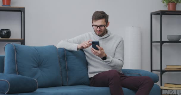 Junger Mann mit Brille surft zu Hause per Smartphone im Internet, sitzt allein auf blauem Sofa im Wohnzimmer, Männerporträt — Stockvideo