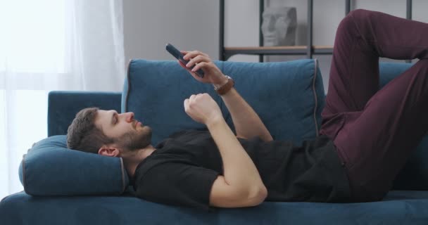 Relaxado cara é navegar na internet e visualização de fita de notícias em redes sociais no smartphone, deitado no sofá no apartamento — Vídeo de Stock