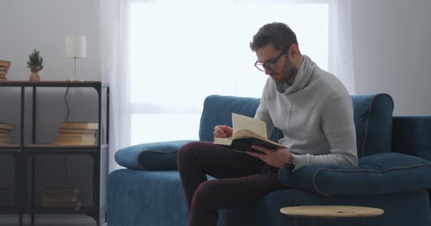 Intelligenter Mann mit Brille liest Buch, sitzt am Wochenende in Wohnung, Isolation und Aussperrung — Stockvideo