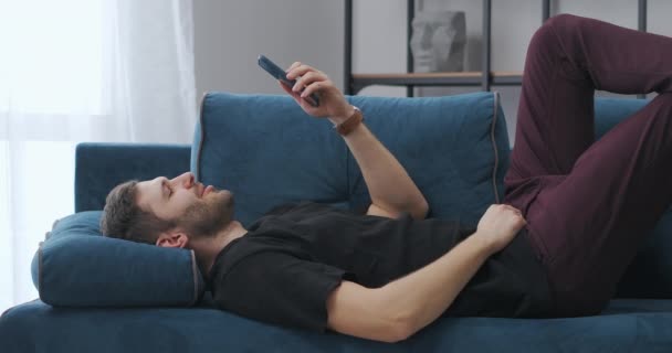 Чоловік весело спілкується за допомогою відео чату в мобільному телефоні, лежить на дивані в квартирі, весело спілкується з друзями або сім'єю, розслабляється — стокове відео