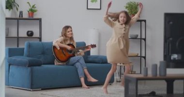 Komik küçük kız, annesi evde gitar çalarken dans ediyor. Mutlu çocukluk ve ebeveynlik, hafta sonunu birlikte geçirmek.