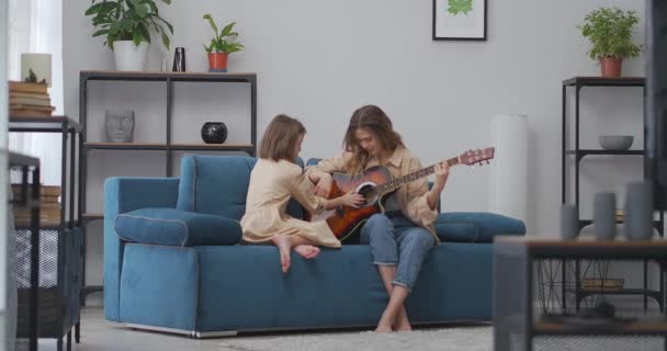 娘とお母さんはリビングで一緒に座ってギターを弾いていて女は楽器を持っていて子供は弦に触れている — ストック動画