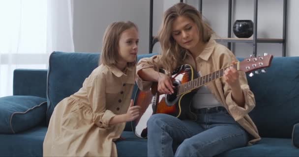 Взрослая женщина и маленькая девочка веселятся вместе дома, леди играет на гитаре, а ребенок разговаривает с ней, семейный уикенд — стоковое видео