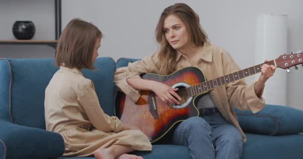 Lección de música de madre a hija en casa, la mujer está tocando la guitarra para niña preescolar, sentados en el sofá juntos — Vídeo de stock