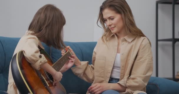 Mulher encantadora e sua filha curiosa estão fazendo música, sentados em casa juntos, menina está tocando guitarra — Vídeo de Stock