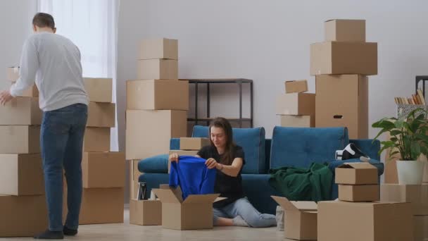 Egy aranyos lány ül a padlón a kanapé mellett, kicsomagol dolgokat, miután elköltözött. Egy karcsú fiatalember kartondobozokat cipel. A család együtt fog élni egy új lakásban.. — Stock videók