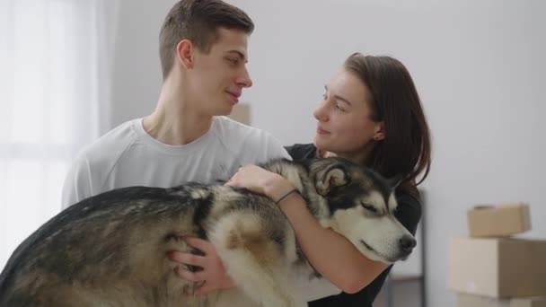 Счастливая молодая супружеская пара держит на руках симпатичную хаски-собаку в новой просторной квартире. Мужчина и женщина сняли новую квартиру. — стоковое видео